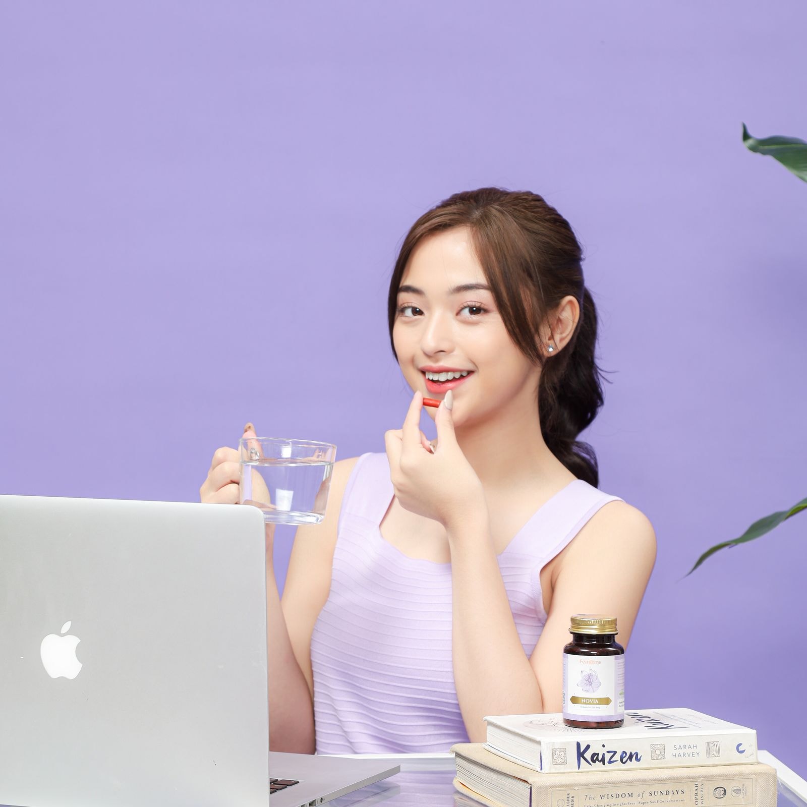 Wanita berbaju lilac memegang kapsul merah dan segelas air, dengan botol Femoire Novia di meja.