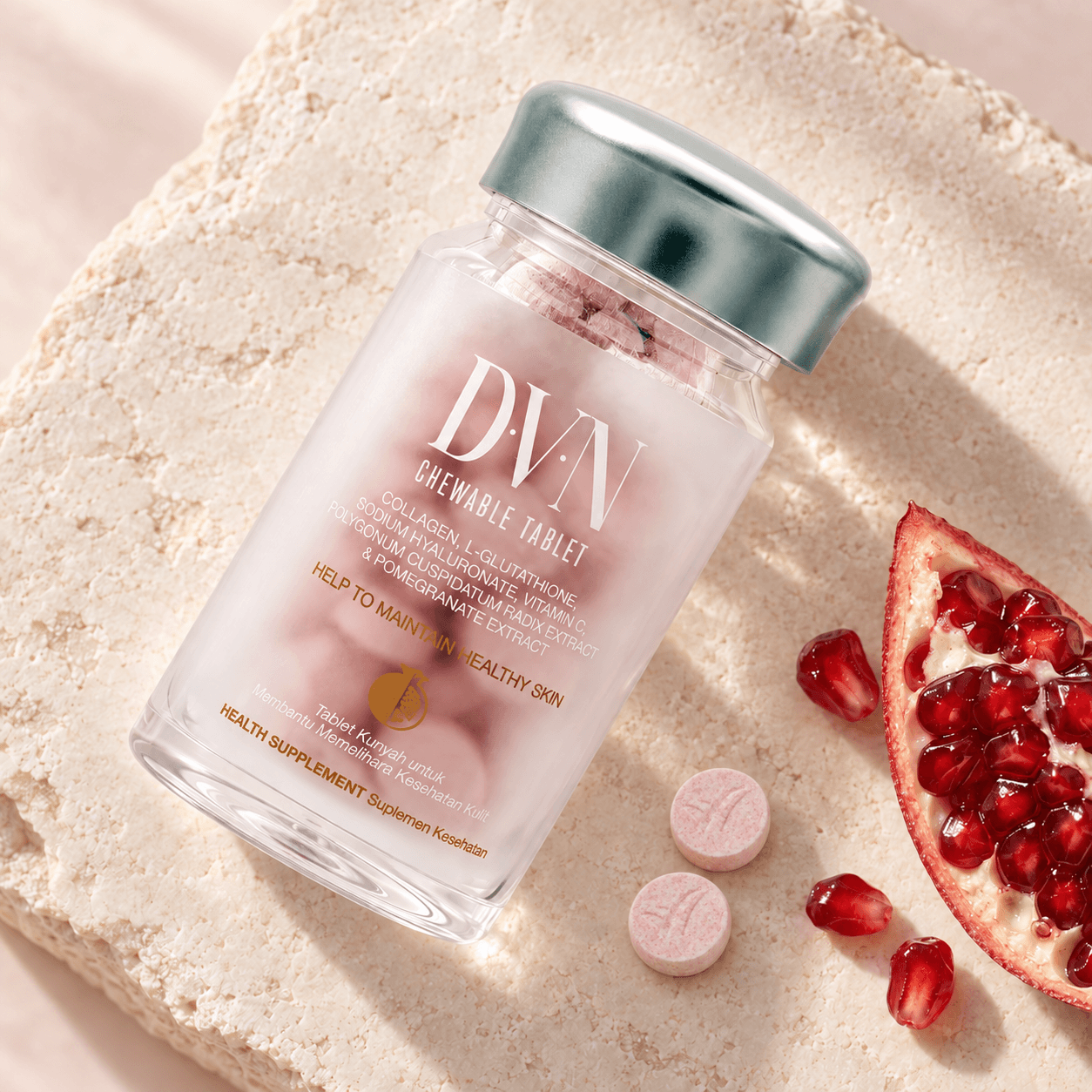 Ilustrasi fotografis botol DVN, dua tablet kunyah tebal berwarna pink krem berbintik dengan logo terukir, dan potongan delima di atas batu ivory.